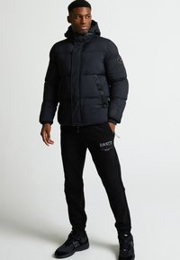 Melns pūkains mētelis ar kapuci, kuram ir kvilts dizains un sānu rāvējslēdzēji. Saskaņots ar melniem jogger tipa biksēm, uz kurām ir "ZAVETT" logo. Melnas sporta apavus.