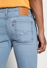 Ljusblå denimjeans har en vävd textur, en brunt läderetikett på midjan och en signatur röd logotag på bakfickan.
