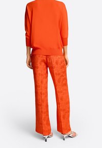 Oranje gebreide trui met driekwartmouwen, gecombineerd met losse oranje broeken met een subtiel bloemenpatroon. Zilveren hakken maken de look compleet.