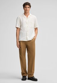 Korte mouwen witte blouse met een textuurpatroon van bloemen, voorzak, gecombineerd met tan rechte broek en zwarte schoenen.