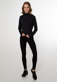 Maglietta nera a maniche lunghe con colletto a zip, abbinata a leggins neri e scarpe sportive nere, posata su uno sfondo neutro.