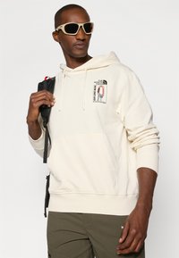 Kremowy sweatshirt z kapturem i kieszenią z przodu, z grafiką i sznurkiem w kapturze. Model nosi beżowe okulary przeciwsłoneczne i czarny plecak.