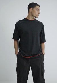 Camiseta negra de manga corta, con un detalle rojo en el cuello y el dobladillo, combinada con pantalones cortos cargo negros holgados. Textura de tela suave.