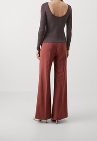 En kvinna står med ryggen mot kameran, iklädd en djupt urklippt, långärmad topp och vida, rostfärgade byxor med klackar.