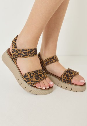 Pieds portant des sandales à semelles épaisses avec des brides en tissu imprimé léopard marron et noir, orteils ouverts et semelles en caoutchouc beige texturé.