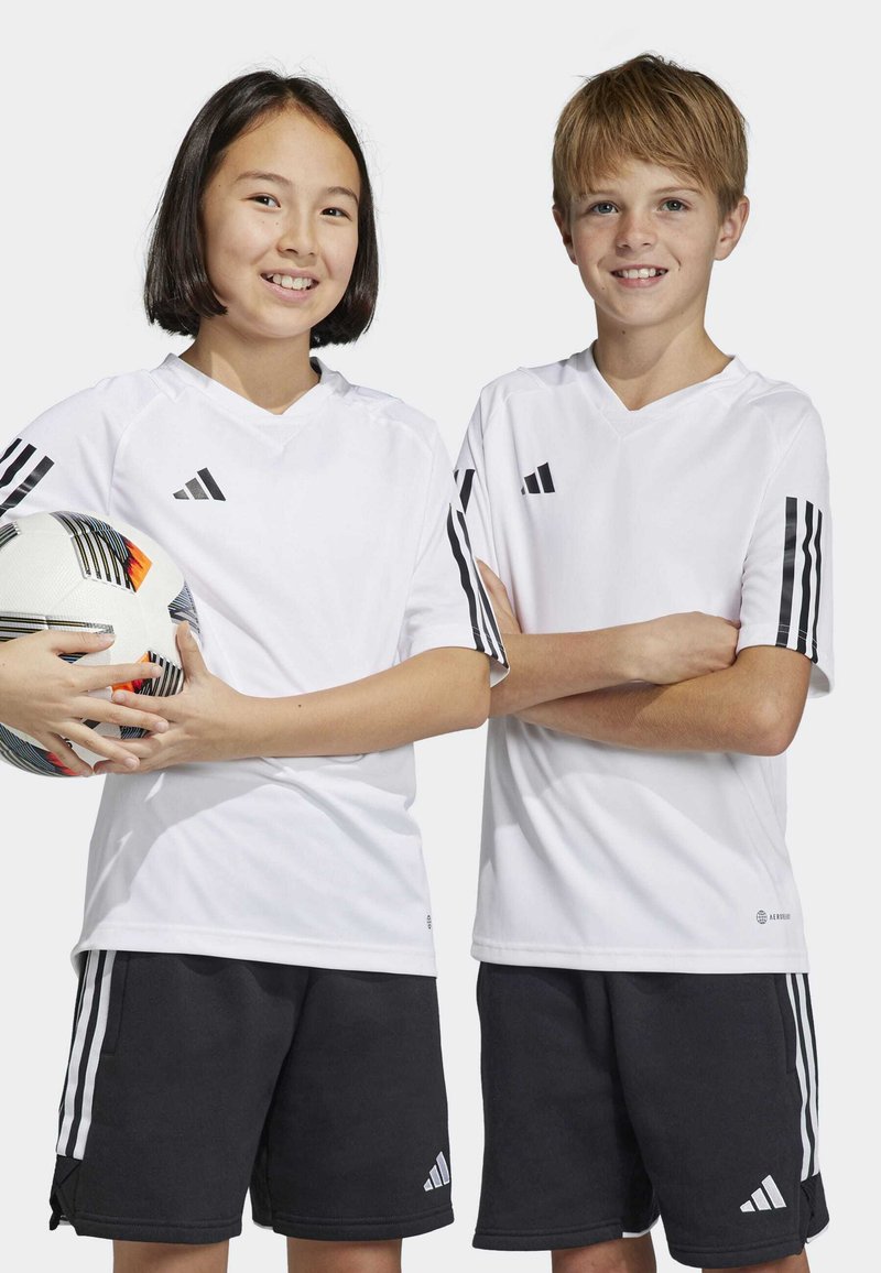 Des t-shirts de sport blancs avec le logo Adidas noir, accompagnés de shorts noirs comportant trois bandes blanches. Une fille tient un ballon de football multicolore.