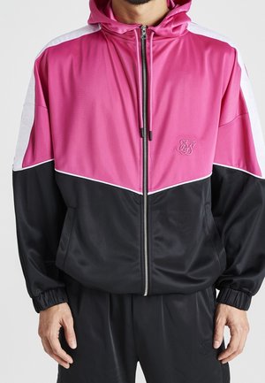 Mand iført en lyserød, sort og hvid hættejakke med lynlås og matchende sorte bukser, stående med hænderne afslappet ved siden.