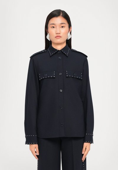Chemise boutonnée navy avec des détails cloutés sur les épaules et les poignets, dotée de deux poches avant et d'un col classique. Texture lisse.