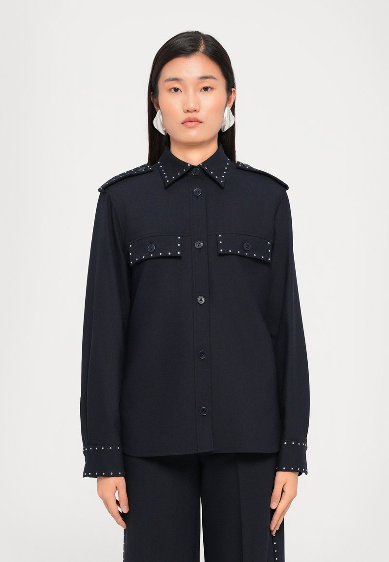 Navy button-up shirt with studded details on shoulders and cuffs, featuring two front pockets and a classic collar. Smooth texture.