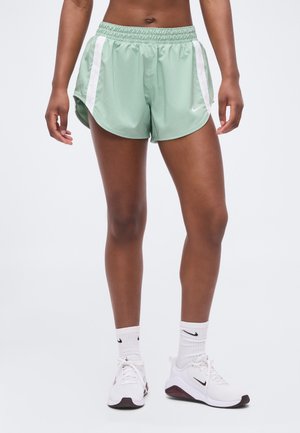 Person trägt hellgrüne Sportshorts mit weißen Streifen, weiße Nike-Sneaker und weiße Nike-Socken und steht vor einem einfarbigen Hintergrund.