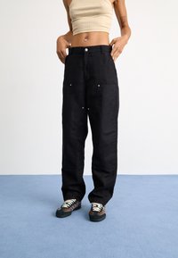 Pantalones anchos negros con bolsillos frontales, herrajes plateados y un corte holgado, combinados con zapatillas multicolores sobre un fondo de moqueta azul.