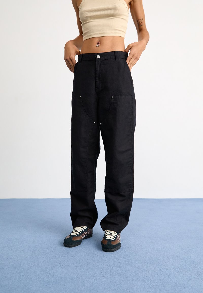 Pantalones anchos negros con bolsillos frontales, herrajes plateados y un corte holgado, combinados con zapatillas multicolores sobre un fondo de moqueta azul.