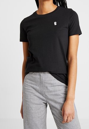 Frau trägt ein schwarzes T-Shirt mit kurzen Ärmeln mit kleinem weißem Geisterlogo und grau karierte hoch taillierte Hose.