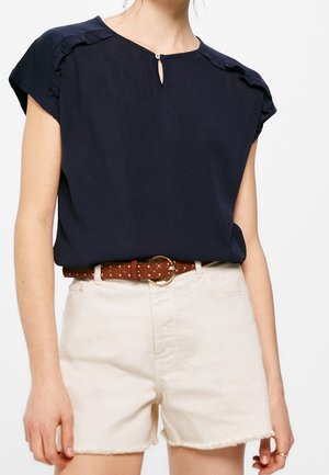 Kobieta ubrana w granatowy, krótki top z wycięciem w kształcie łezki oraz beżowe, wysokie szorty z brązowym paskiem ozdobionym ćwiekami.
