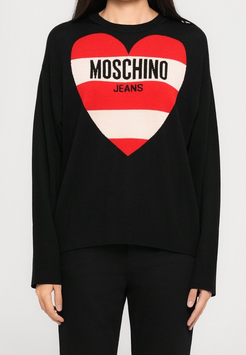 Personne portant un pull noir à manches longues avec un grand cœur rouge et blanc sur le devant et le texte "MOSCHINO JEANS" à l'intérieur du cœur.