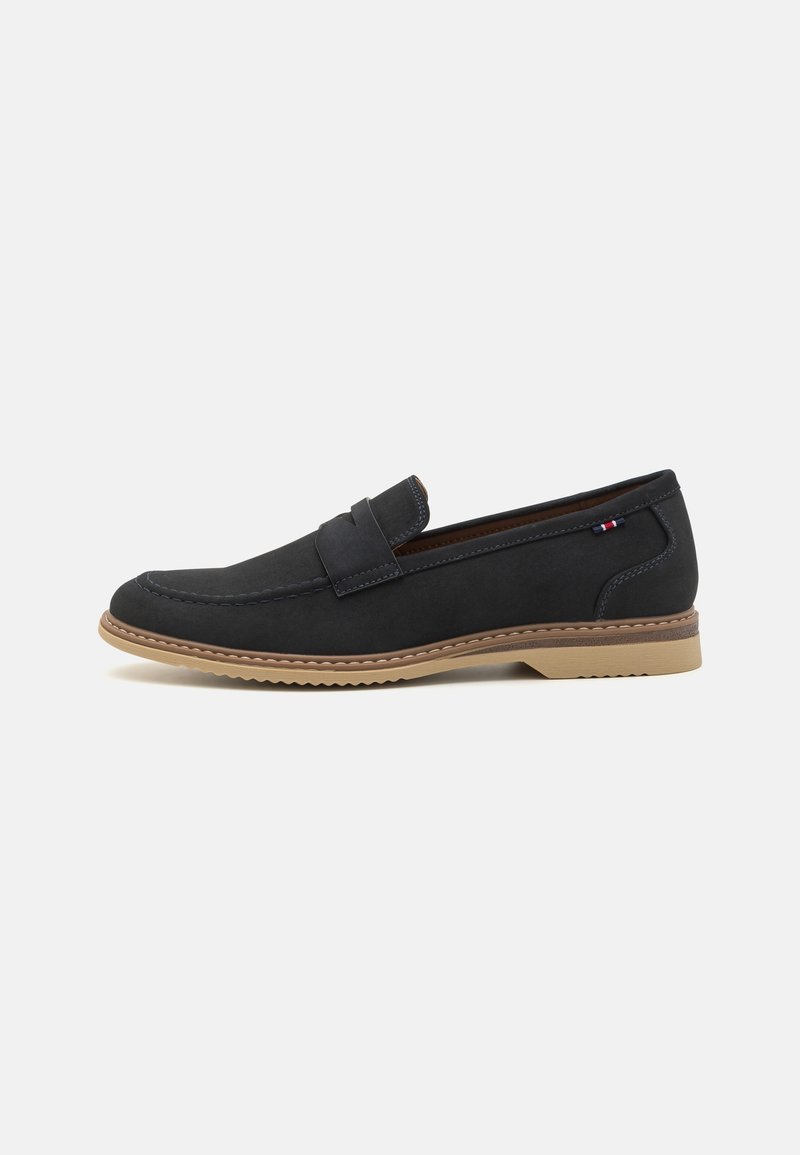 Mocassin noir en cuir à enfiler avec semelle en caoutchouc couleur noisette et petit écusson en tissu logo sur le côté du talon, vue de profil latéral sur fond blanc.