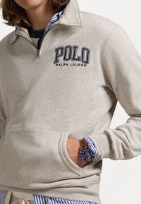 Grå sweatshirt med halvdragkedja, med en "POLO"-logotyp i marinblått. Inkluderar en ficka fram och en lager av blårandig skjorta under.