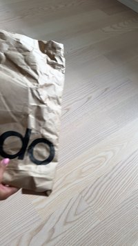 Guban rjav papirnati dodatek z velikim črnim tiskom "do", drži se s strani, počiva na svetlem lesenem tlehu z vidno strukturo lesa.
