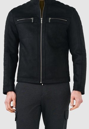 Hombre con chaqueta negra con cremallera, dos bolsillos horizontales en el pecho y pantalones gris oscuro sobre un fondo liso.