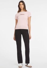 Camiseta de algodón rosa claro con mangas cortas, que presenta el logo negro "GUESS JEANS" en el frente. Combinada con jeans negros ajustados y zapatillas deportivas.