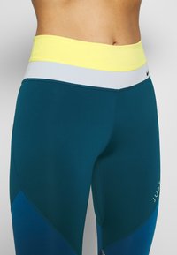 Leggings high-waisted color teal con cintura elastica gialla e grigia, caratterizzati da una texture liscia e un design geometrico a blocchi di colore.