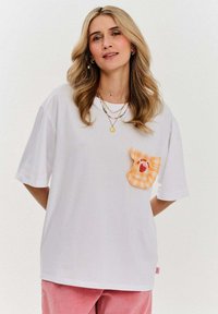 Bílá tričko s krátkým rukávem, zdobené oranžovou kostkovanou kapsou s designem jahody. V kombinaci s růžovými manšestrovými kalhotami.