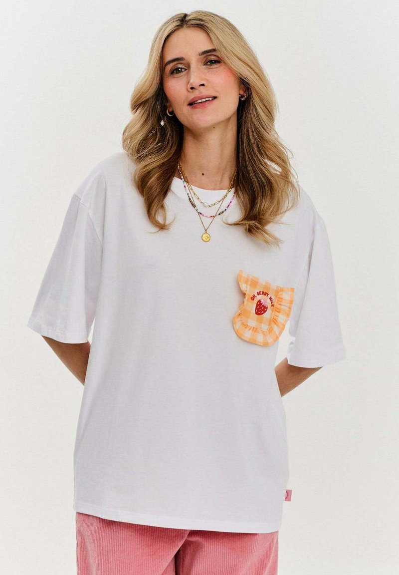 Bílá tričko s krátkým rukávem, zdobené oranžovou kostkovanou kapsou s designem jahody. V kombinaci s růžovými manšestrovými kalhotami.