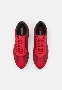 Zapatillas deportivas rojas con parte superior de malla texturizada y acentos sintéticos lisos. Presentan cordones planos y plantillas negras con marca visible.