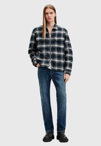 Chaqueta de franela a cuadros en negro y gris con cremallera, dos bolsillos en el pecho, combinada con jeans de mezclilla azul y botas negras hasta el tobillo.