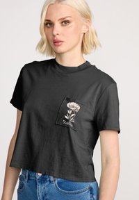 Czarny krótkie t-shirt z miękkiego materiału z okrągłym dekoltem. Posiada kieszonkę z kwiatowym wzorem i tekstem na szwie.