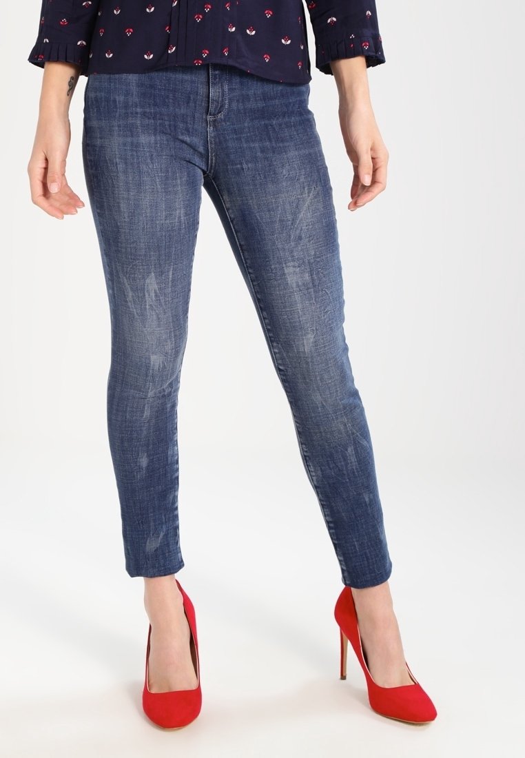 Femme portant un jean skinny bleu délavé et des chaussures à talons rouges, debout avec les mains relâchées le long du corps, sur un fond blanc uni.