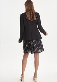 Czarna marynarka o strukturze, zestawiona z prześwitującą czarną spódnicą w groszki. Strój cechuje warstwowa kompozycja i lekka tekstura materiału.