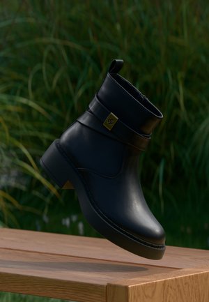 Black leather ankle boot with a decorative strap and gold emblem, hovering above a wooden surface with green foliage background.