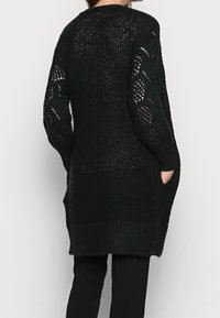 Svart stickad cardigan med lös väv, långa ärmar och sidofickor. Ribbad nederkant och ärmslut; texturerat mönster på ärmarna.