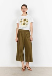 T-shirt bianco con stampa floreale dorata e testo, abbinato a culottes plissettate verde oliva e mules con tacco marroni su uno sfondo neutro.