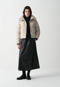 Beige pufferjacka med en strukturerad passform och texturerad mönster, kombinerad med en svart lärokjol och svarta skor, framför en ensartad bakgrund.