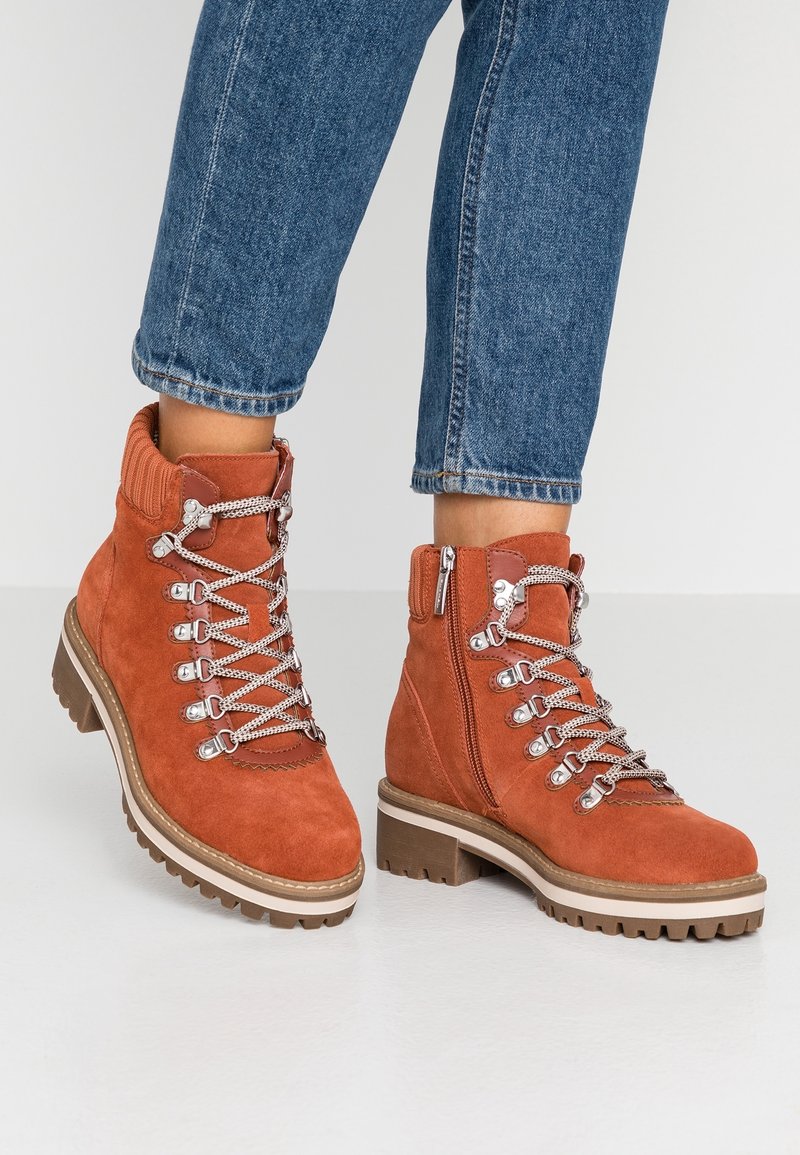 Bottines en daim couleur rouille avec semelles à crampons épaisses, lacets contrastés, fermetures éclair latérales, portées avec un jean bleu retroussé sur fond blanc.