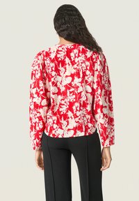 Rød blomstret bluse med hvide mønstre, lange ærmer, rund halsudskæring og sammenfoldede detaljer ved skuldrene; parret med sorte bukser.