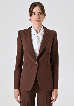 Blazer marrone su misura con un unico bottone dorato, rever a scollo e due tasche anteriori, abbinato a una blusa bianca con collo a cravatta.