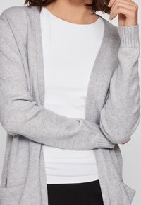 Jasnoszary kardigan wykonany z miękkiego dzianinowego materiału, z otwartym przodem, ściągaczami na mankietach i kieszeniami z przodu, noszony na białym topie.