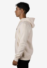 Sudadera con capucha color crema hecha de tela suave con un bolsillo tipo canguro y cordón. Cuenta con puños y dobladillo acanalados; se lleva con pantalones oscuros.