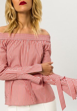 Kobieta nosząca czerwoną i białą bluzkę z pionowymi paskami, odsłaniającą ramiona, z długimi rękawami związanymi na nadgarstkach, w połączeniu z białymi spodniami.