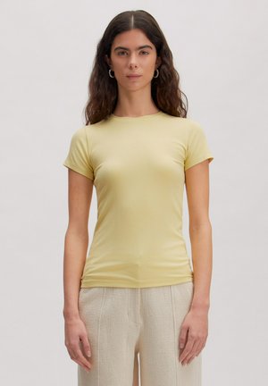 Vrouw met lang donker haar, gekleed in een aansluitend lichtgeel topje met korte mouwen en crème getextureerde broek, staand tegen een effen achtergrond.