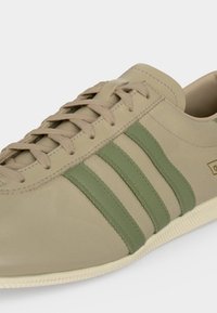 Basket en cuir beige avec des rayures vert olive, des lacets beiges et une semelle blanche, montrée en gros plan de côté.