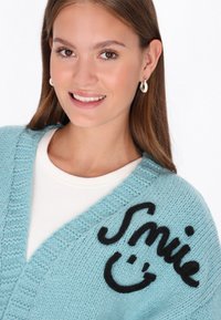 Ljusblå stickad kofta med V-hals, med en svart broderad "Smile" och en glad ansiktsdesign på framsidan. Mjuk textur.