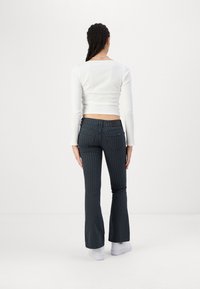 Mujer con el cabello trenzado de espaldas, vistiendo una camiseta blanca de manga larga, jeans de campana a rayas negras y zapatillas blancas, sobre un fondo blanco.