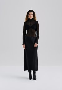 Svart strikket turtleneck med gjennomsiktig stripete design, kombinert med et langt svart tettsittende skjørt og svarte hælboots. Jevn tekstur gjennom hele.