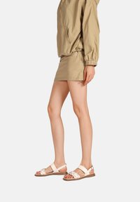 Giacca leggera beige, gonna abbinata e sandali chiari con accenti metallici, suola piatta e dettagli decorativi sulle cinghie.