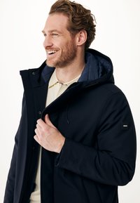 Veste imperméable marine avec capuche, fermeture éclair avant et boutons-pression. Comprend une doublure intérieure douce et un patch de logo sur la manche.