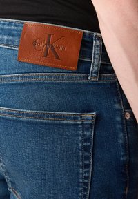 Közeli kép kék farmernadrág hátsó zsebével, barna Calvin Klein bőr márkajelzéssel és egy személy karjával oldalról.